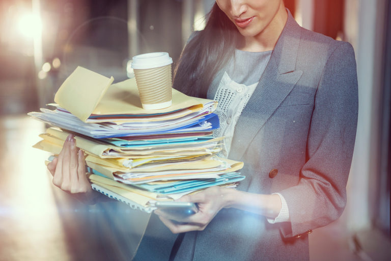 Businesswoman carrying stack of file folders while using mobile